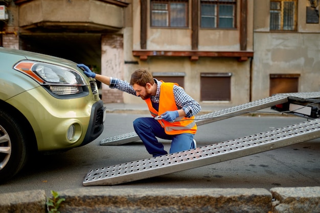 Male road worker preparing car for evacuation on tow truck fasten hook working on city street