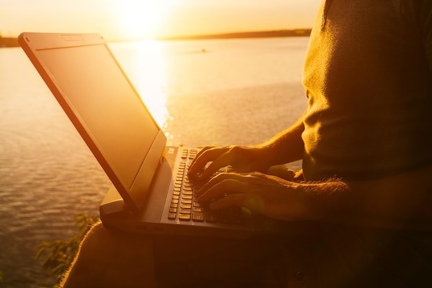 Male's hands working on a laptop in the evening near the river at sunset Work during vacation outdoors in summer Closeup