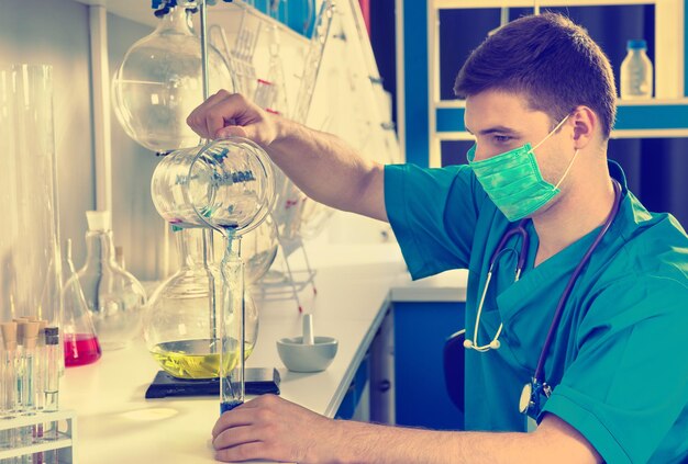 Scienziato maschio in uniforme che indossa una maschera tiene in mano un bicchiere di prova e riempie una provetta con liquido blu, facendo alcune ricerche in laboratorio. concetto di sanità e biotecnologia