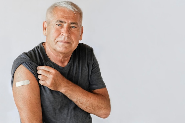 COVID19またはサル痘コビッドからのワクチン接種後に包帯パッチで腕を示す老人灰色のスタジオの背景のためのワクチン接種後に笑顔の男性シニア