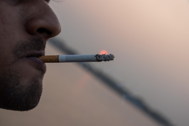 Male smoker with a beard Unhealthy lifestyle with a male smoker Smoker with a cigarette in his mouth on a blurry sunset background Cigarette burning in a smoker's mouth Man smoking closeup view