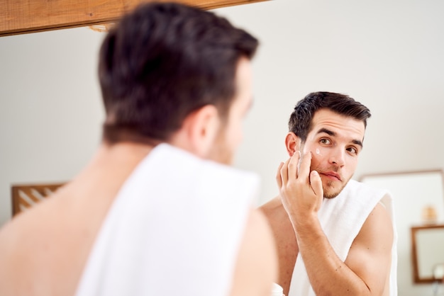 Male spreads his face with white cream while standing in front of mirror in bath at morning