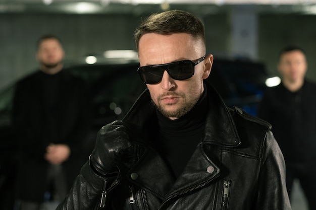 Male spy or gangster in sunglasses, black leather gloves and jacket and his gang or bodyguards on background