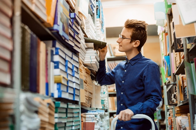 Studente maschio in cerca del libro necessario su numerosi scaffali della biblioteca