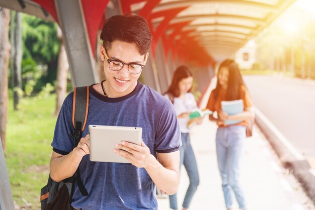 Male student reads on tablet computer at campus. There are groups of girl riends behind, Education concept