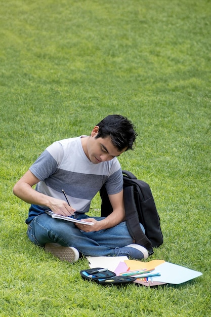 Студент-мужчина сидит на траве и пишет в блокноте.