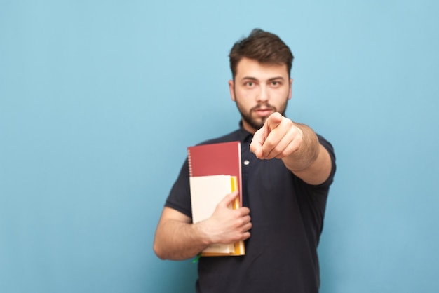 Male student with books in his hands stands on blue and shows the index finger wearing a black shirt