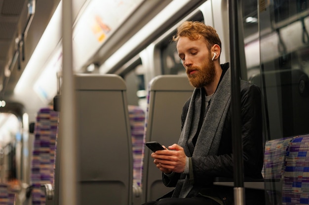 Male subway passenger sits on a train listening to music on headphones and using a smartphone