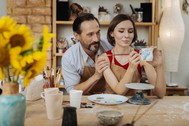 Male support. Bearded man feeling very helpful while supporting his wife working with clay pot