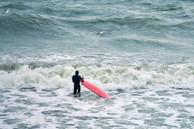 Male surfer in black swimsuit in ocean with red surfboard\
waiting for big wave. warm day, beautiful sea waters, nature scene.\
sport and outdoor activity concept.