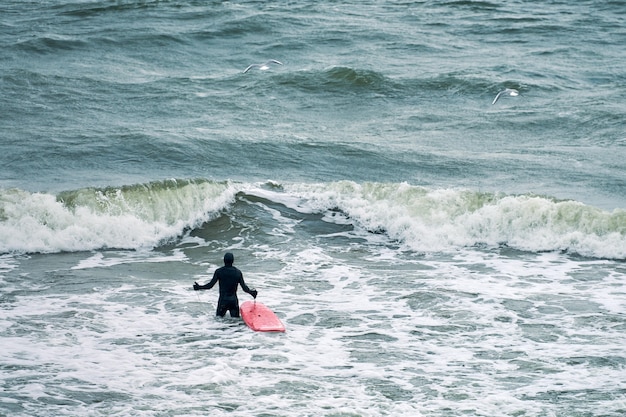 Male surfer in black swimsuit in ocean with red surfboard waiting for big wave