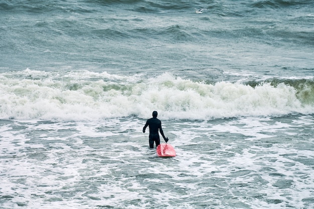 Male surfer in black swimsuit in sea with red surfboard waiting for big wave.