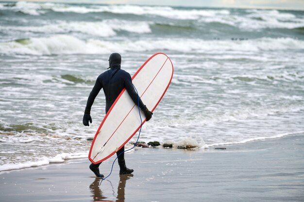 Male surfer in black swimsuit walking along sea and holding white surfboard in his hand