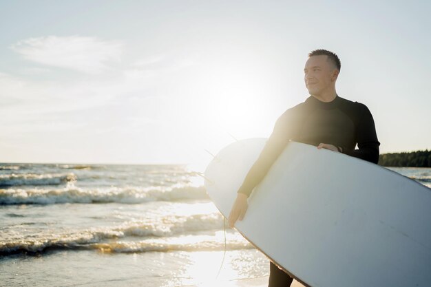 A male surfer in a wetsuit riding in the sea on a sunny day at\
sunset holding surfing in his hands