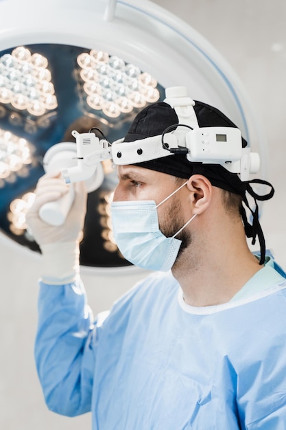 男性外科医は手術室で光ランプを保持し、それを手術野に向ける ヘッドライトを持つ外科医は診療所で手術の準備をしている