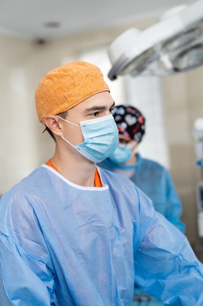 Male surgeon wearing protective cap and mask standing with his colleagues at the background and looking away Medic during operation Medicine concept