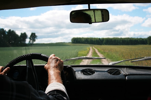 Male tanned hands with a tucked shirt on the steering wheel of a riding car and the road, clouds, forest and field in front.