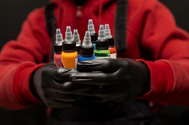 さまざまな色のタトゥーインクのボトルを保持している男性のタトゥーアーティスト黒い手袋と赤いセーターを着て瓶の黒い背景に選択的な焦点ボディアートコンセプト