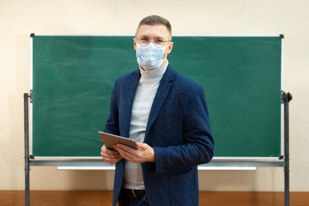 A male teacher in a medical mask stands at the blackboard in the classroom