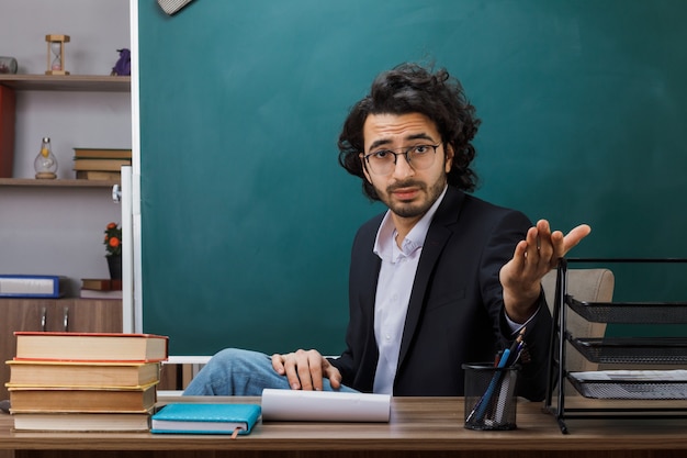 Insegnante maschio con gli occhiali seduto al tavolo con gli strumenti della scuola in classe