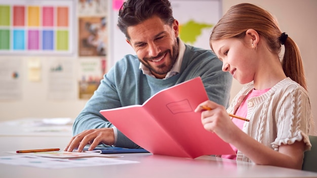 Male Teacher With Female Student In School Classroom Sitting At Desk Writing In Book Together