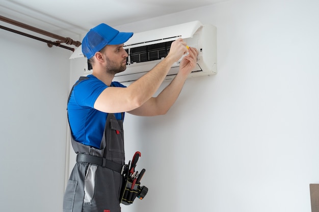 Foto tecnico maschio in tuta e berretto blu ripara un condizionatore d'aria sul muro