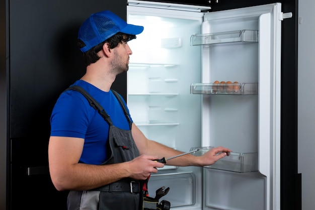 male technician in uniform repairs a broken refrigerator in the kitchen