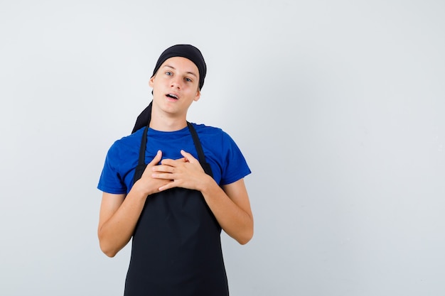 Male teen cook keeping hands on chest, opening mouth in t-shirt, apron and looking surprised. front view.
