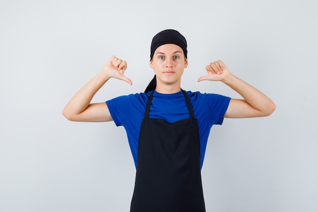 남자 10대 요리사는 티셔츠, 앞치마를 입은 엄지손가락으로 자신을 가리키며 자신감을 보입니다. 전면보기.