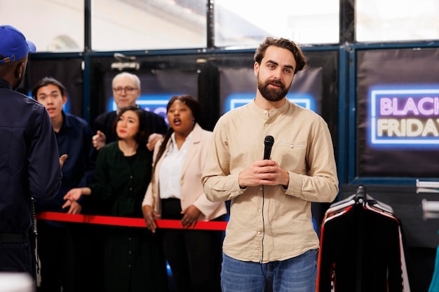 Male television reporter standing in crowded clothing store holding microphone producing live newscast about Black Friday madness. Retail store owner launching start of seasonal sales