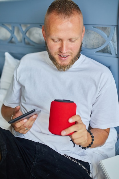 Male testing talking smart wireless speaker, man controlling home devices with a voice commands, sitting on sofa at home, concept of smart home