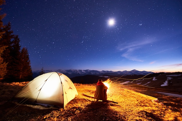 男性の観光客は夜にキャンプファイヤーの近くのキャンプで休憩し、星と月でいっぱいの美しい夜空の下でテントを張り、山の夜景を楽しんでいます。