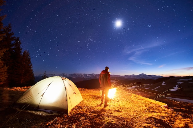 男性の観光客は夜に彼のキャンプで休憩し、星と月でいっぱいの美しい夜空の下でキャンプファイヤーとテントの近くに立って、山で夜のシーンを楽しんでいます