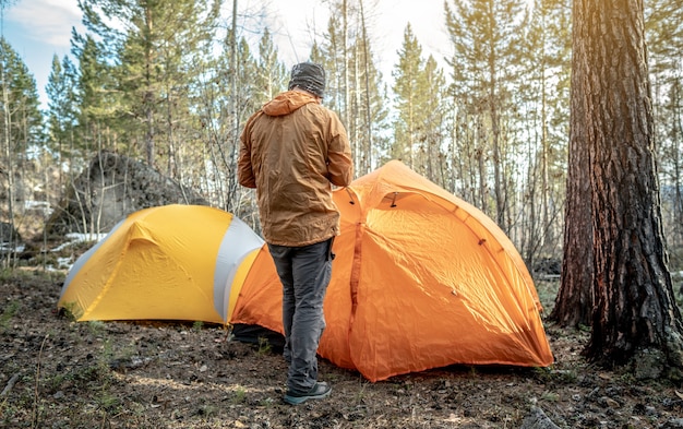 A male traveler is standing in the forest near the tents in a tent camp. Concept of hiking, spending time in nature.