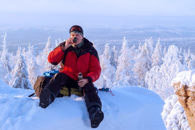 Male traveler sits resting on a cliff in the winter mountains and drinks tea from a thermos