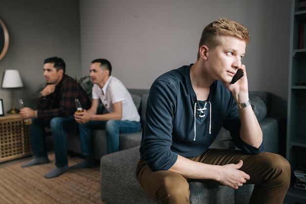 Male trying to talk on the phone Behind the guy back his friends are watching TV Group of friends watching a football match on TV against the background of young man phone conversation