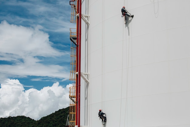 Male two workers control swing rope down height tank rope access inspection of thickness pipeline and tank gas in the blue sky.