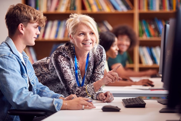 Male University Or College Student Working At Computer In Library Being Helped By Tutor