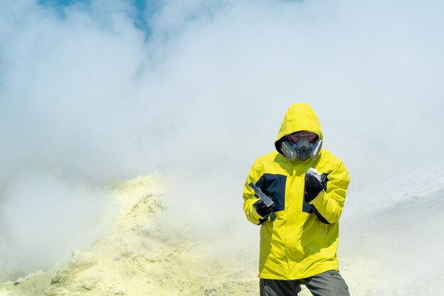 噴気孔の蒸気の中で火山の斜面にいる男性の火山科学者が、鉱物のサンプルを調べる