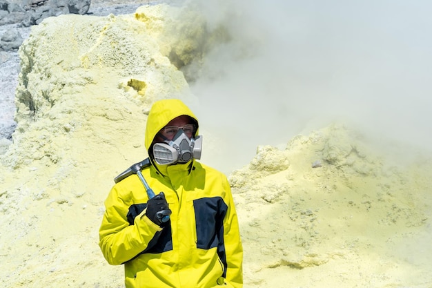 Male volcano scientist with a geological hammer and in a respirator on the slope of a volcano nearby among toxic fumes