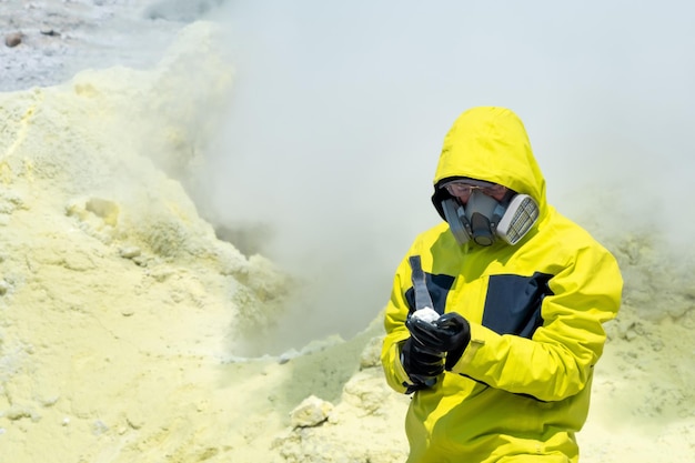 Male volcanologist on the background of a smoking fumarole examines a sample of a sulfur mineral with a geological hammer