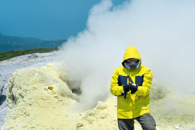 噴気孔を背景に男性の火山学者が地質ハンマーで硫黄鉱物のサンプルを調べる