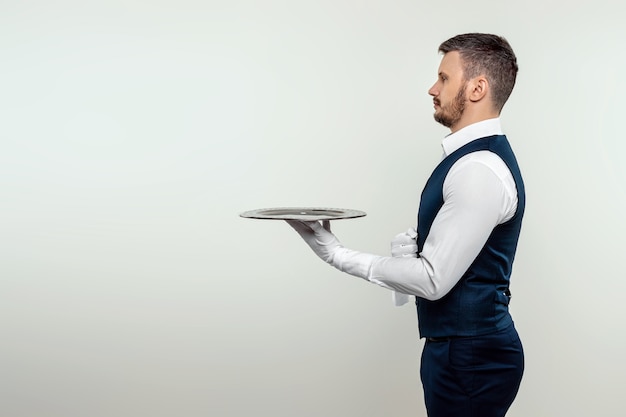 A male waiter in a white shirt stands sideways with a silver tray. The concept of service personnel serving customers in a restaurant.