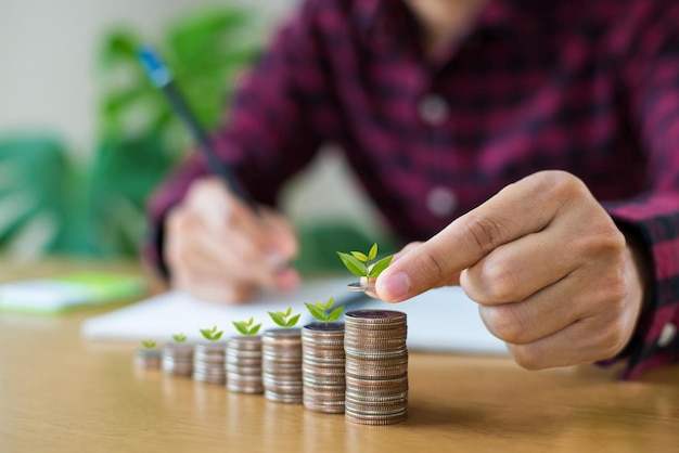 Male wearing plaid shirt hand putting money coins stack growing business with tree growing on money on desk with copy space