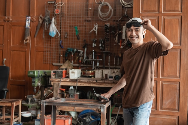 Male welder smiles at the camera holding sunglasses and welder against a welding workshop background