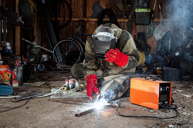Мужской сварщик в сварочной маске работает с дуговым электродом в своем гараже. Сварка, строительство, металлообработка.