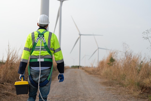 Photo behind male windmill engineer wear uniform with helmet safety hold tool box and walking to wind farm