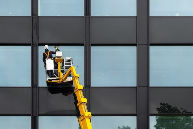 Фото Мужчина мойщик окон чистит стеклянные окна на современном здании высоко в воздухе на платформе лифта. рабочий полирует стекло высоко в воздухе