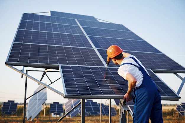 Male worker in blue uniform outdoors with solar batteries at sunny day.