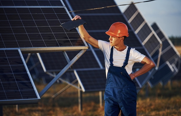 Male worker in blue uniform outdoors with solar batteries at sunny day.